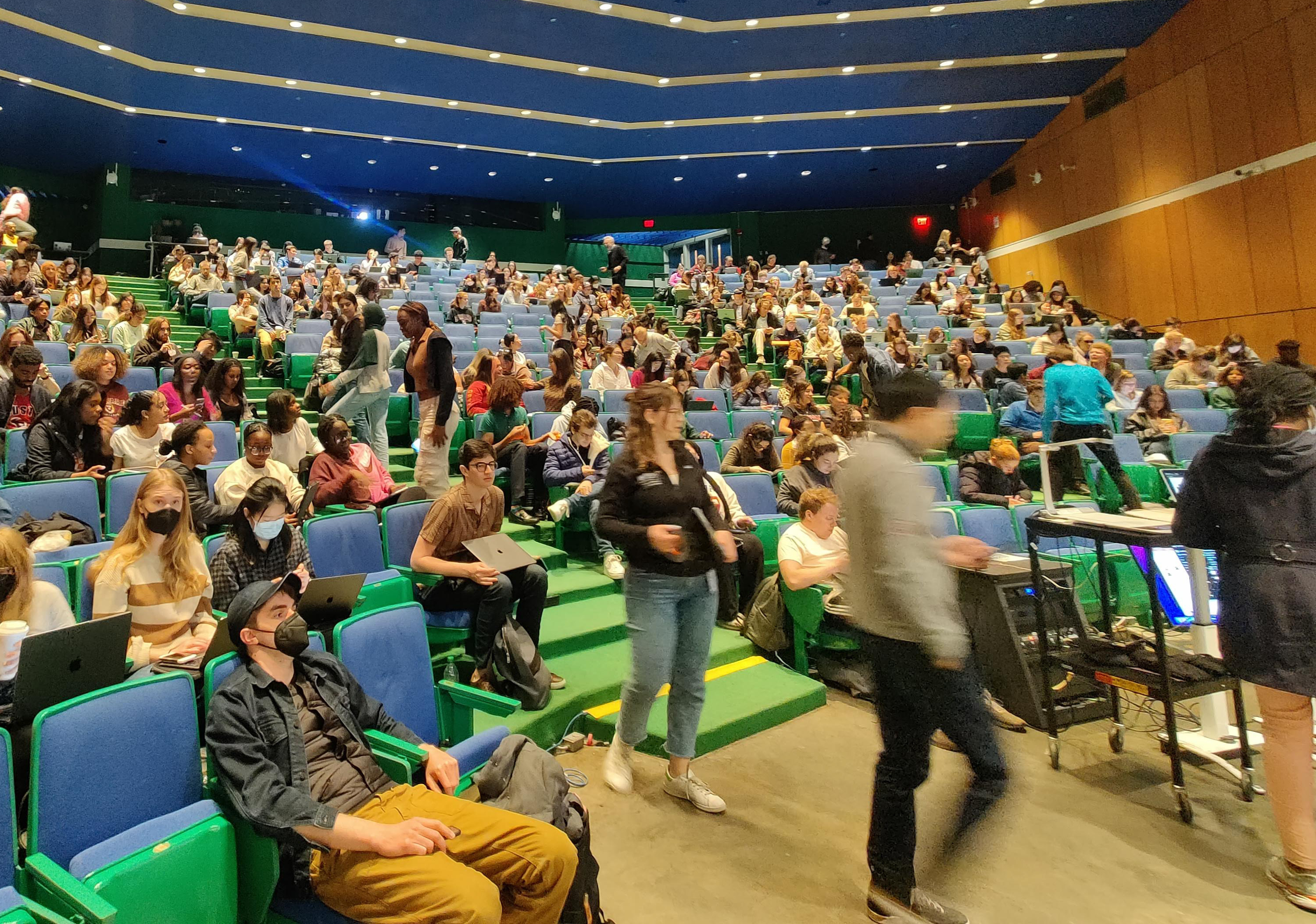 A large lecture hall with blue seats and green carpeting on each tier. Lots of movement going on: some students are still filing in, while others are sitting with their laptops or talking to each other.