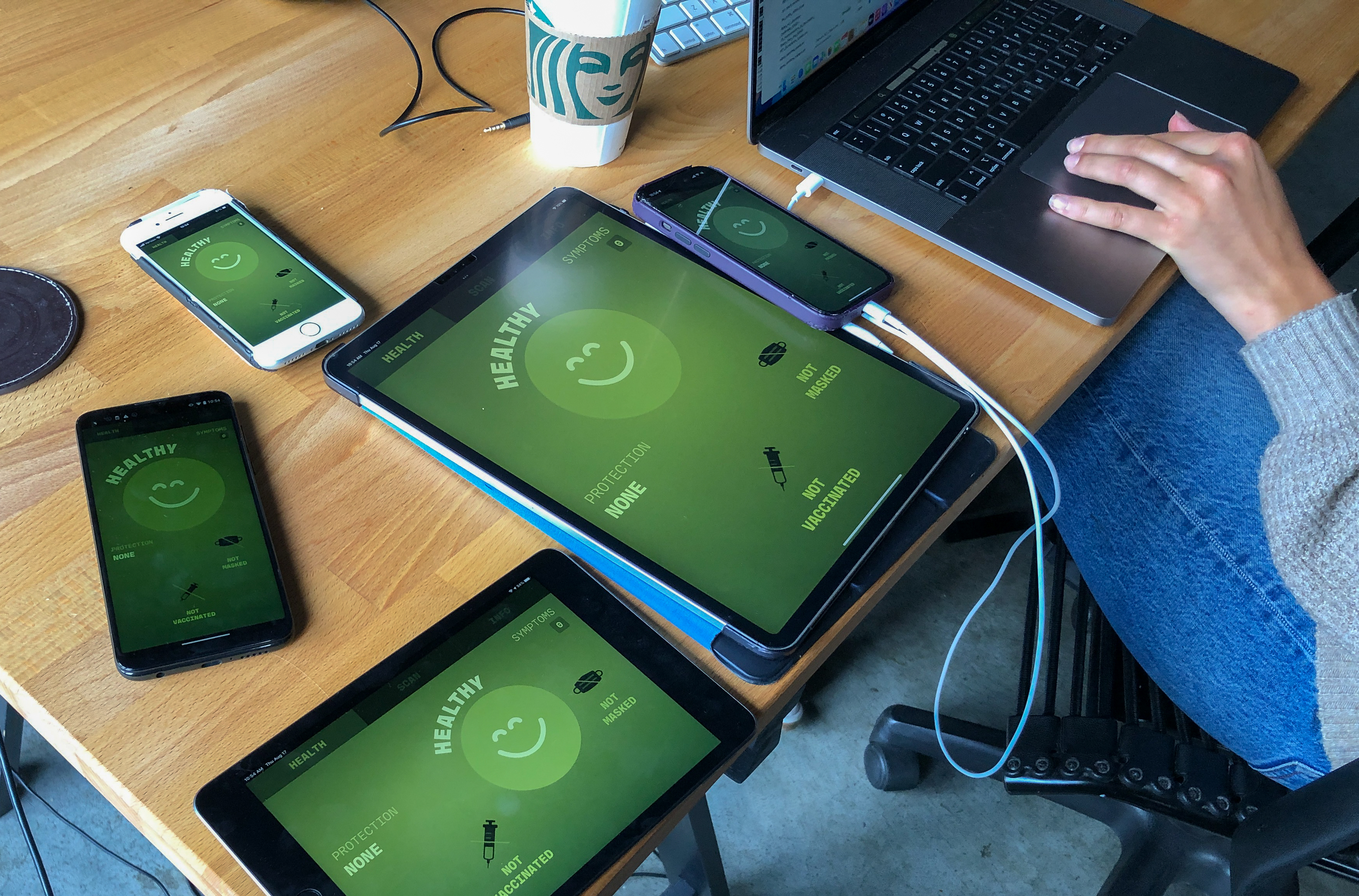 Five different devices all laid out on the desk, each showing the Operation Outbreak mobile screen. A smiley face on a green background represents the participant’s healthy status, and the screen also shows that the participant currently has no protection, is not vaccinated, and is not masked.