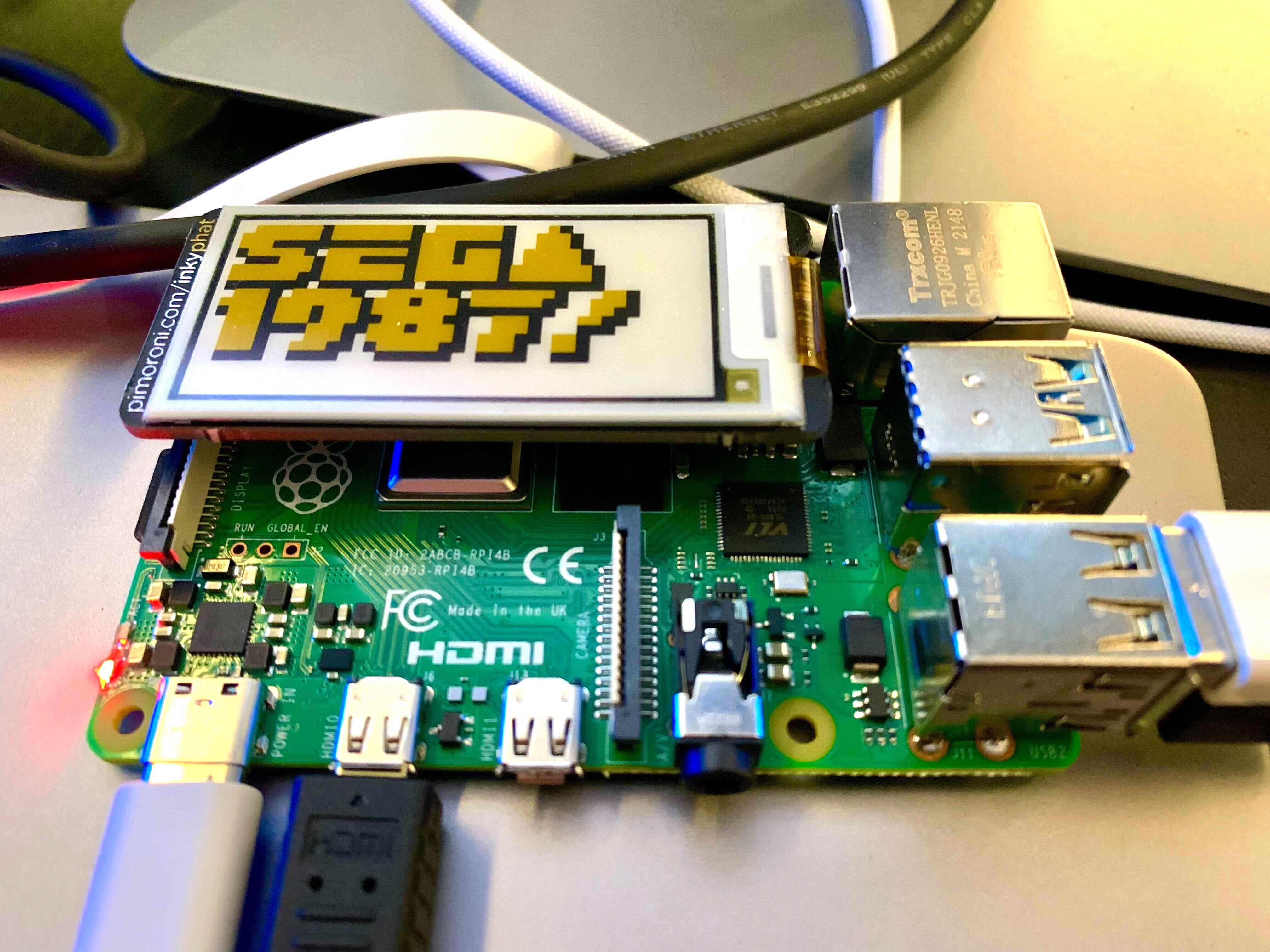 A small e-ink screen on a Raspberry Pi, displaying the words “SEGA 1987!” in a smooth and crisp retro video game typeface, in yellow with a black shadow.