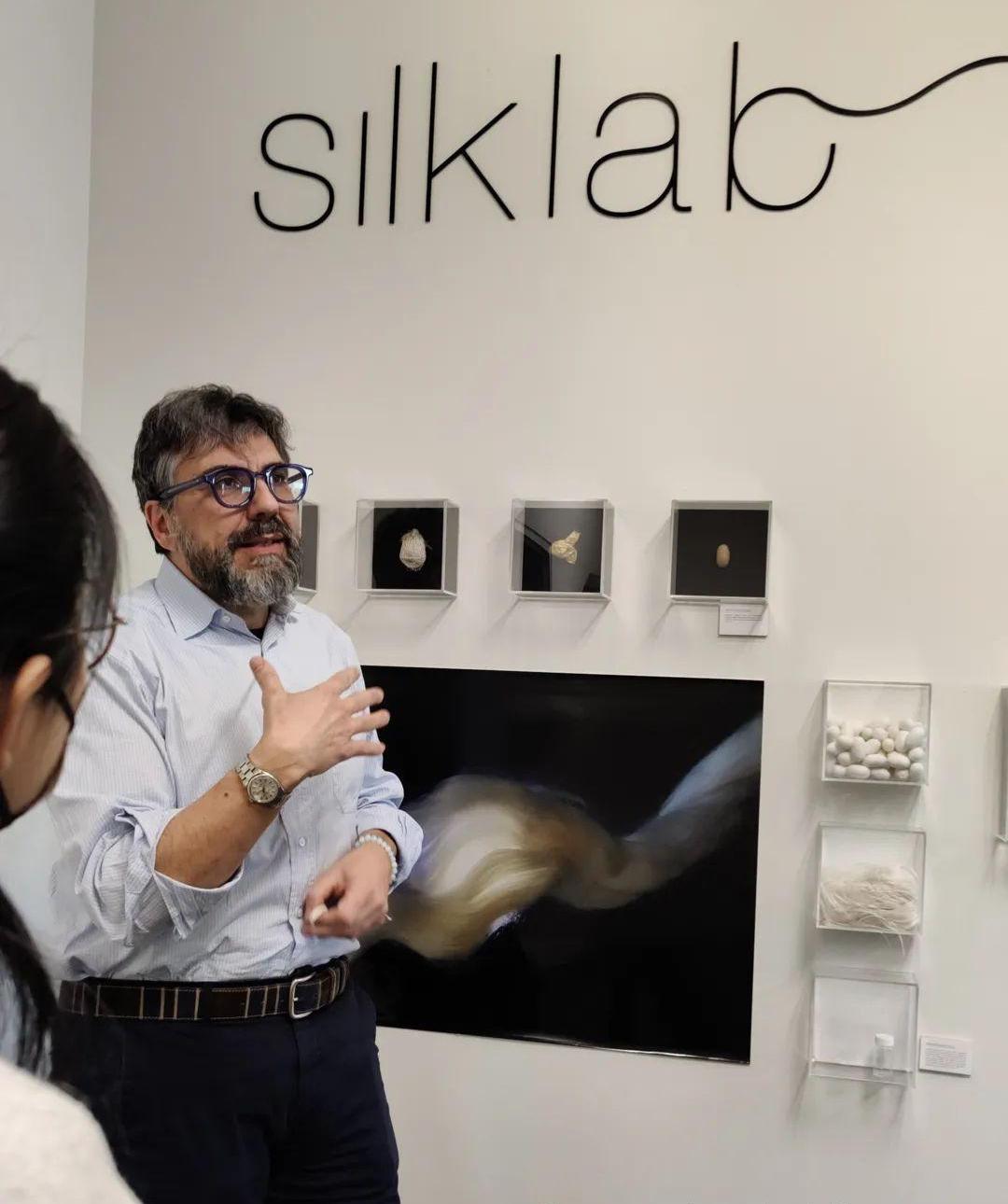 Fio Omenetto stands in front of one of the wall displays at Silklab, mid-explanation. Behind him are small square display cases that house examples of the silk at different stages of processing.