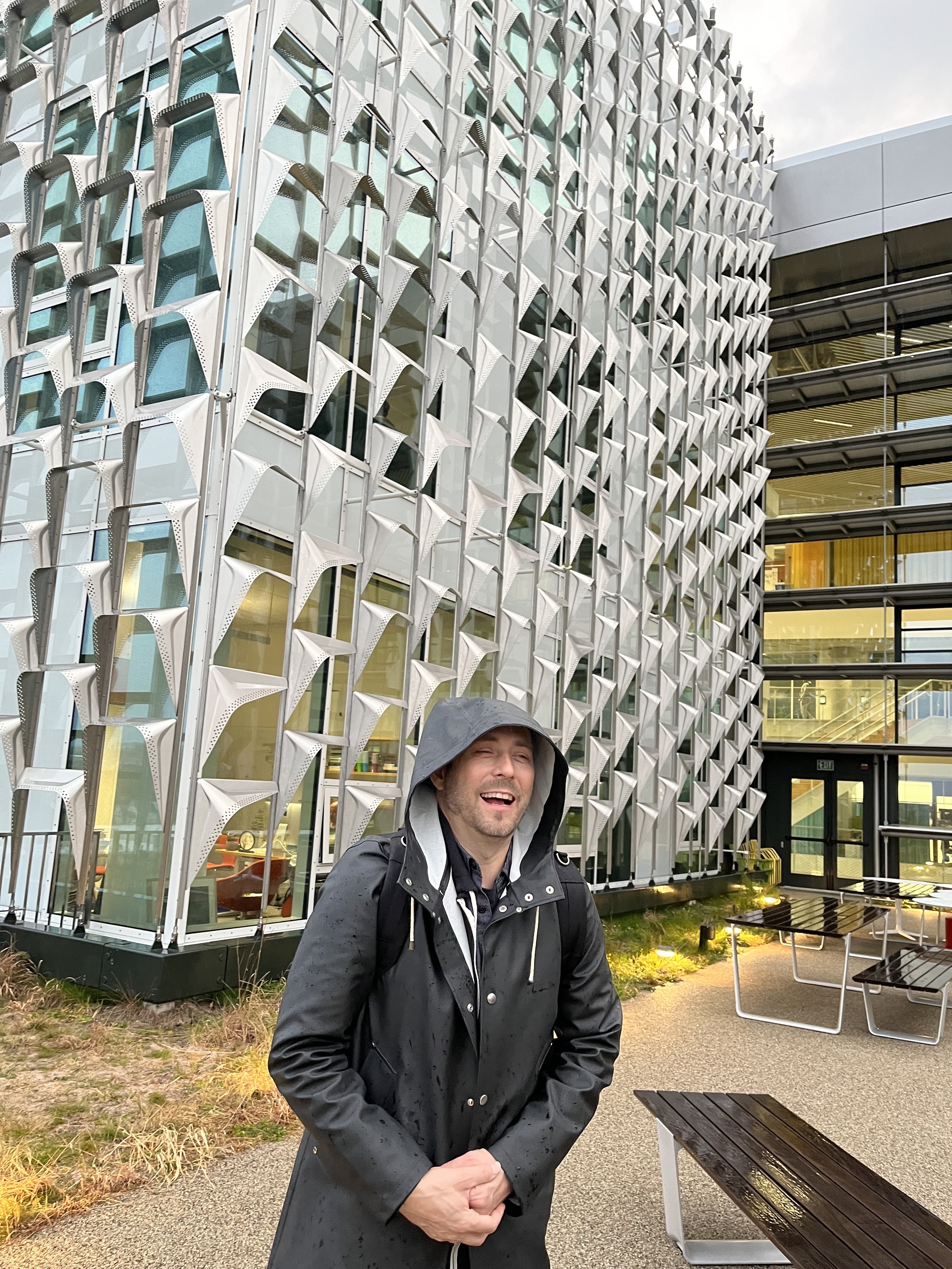 Tim stands in a black raincoat outside in front of a glass building with decorative metal window shades jutting out every few feet to form an intricate grid pattern.