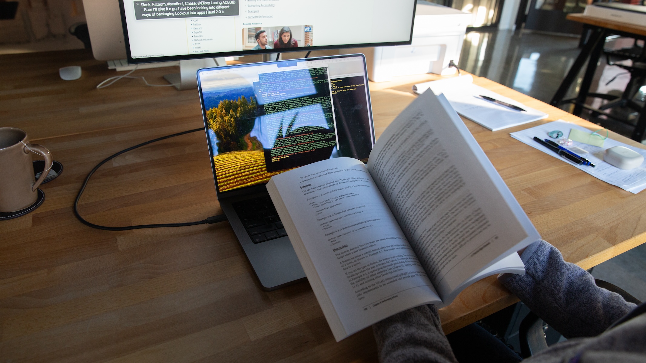 A person sitting at her desk, flipping through a book about accessibility while looking at a terminal on a laptop screen