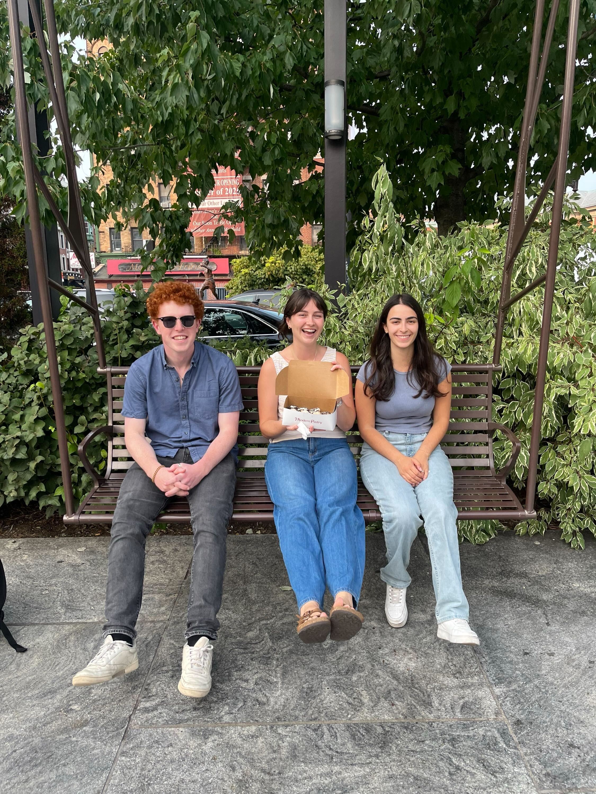 Andy, Laura, and Elissa sit together on a swing in the North End. Laura holds a box of cannoli and looks very excited about it.