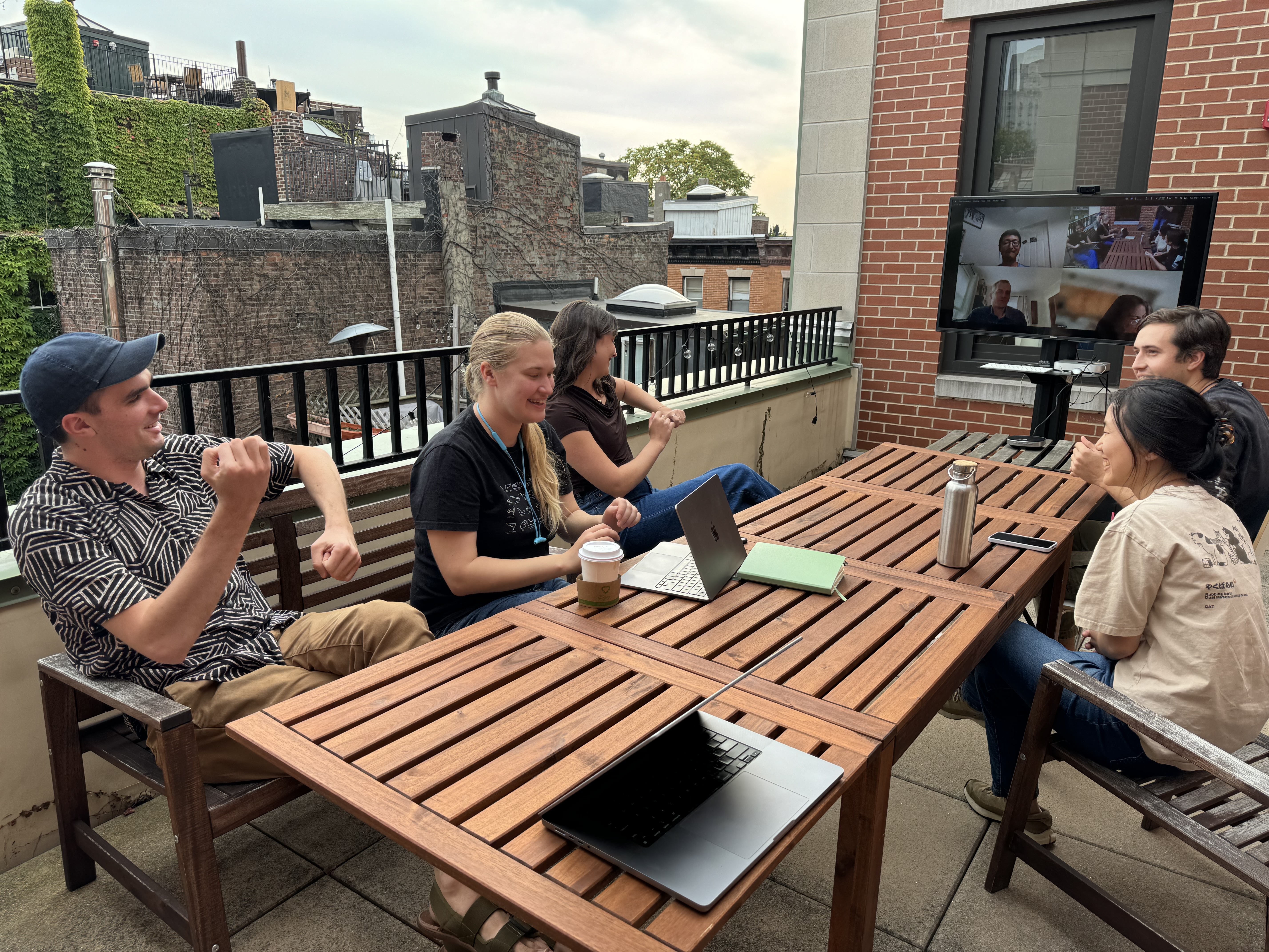 Five people sit around a patio table and three others have joined via Zoom. They are laughing and making rowing motions with their arms in celebration of the Rowboat launch.