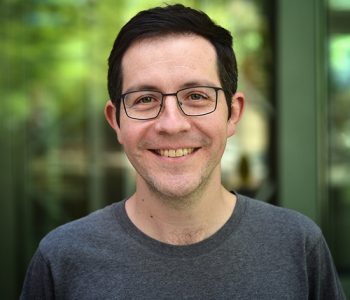 Headshot of our collaborator Patrick wearing a grey shirt standing in front of a blurred green background. 