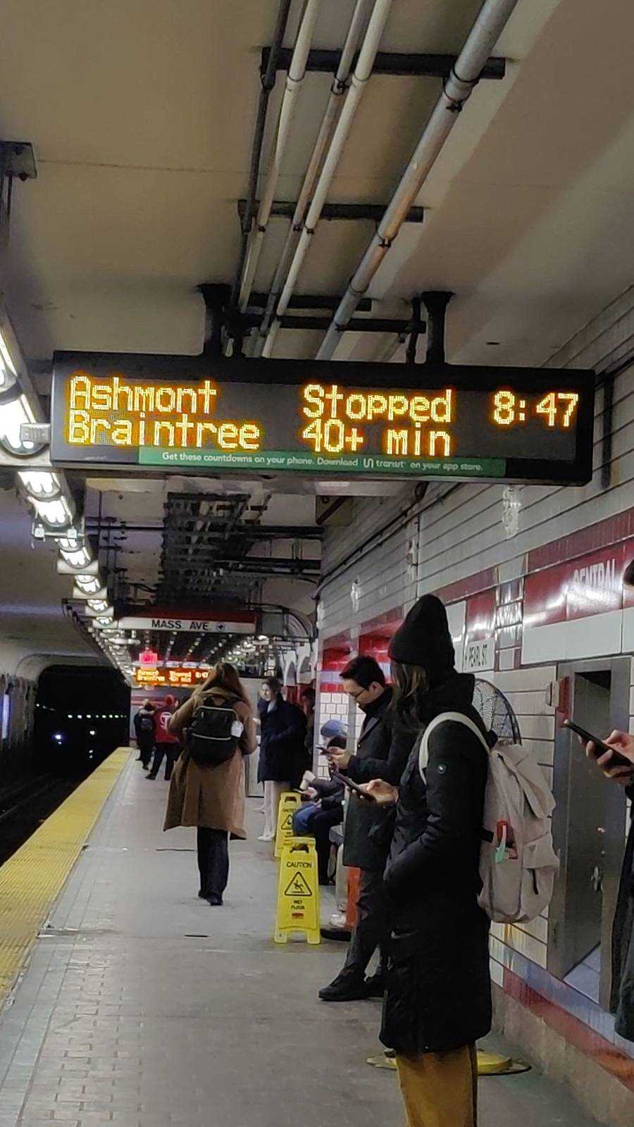 Several commuters, bundled in their winter coats, stand under a sign telling them their train is stopped and it will be 40+ minutes until the next one.